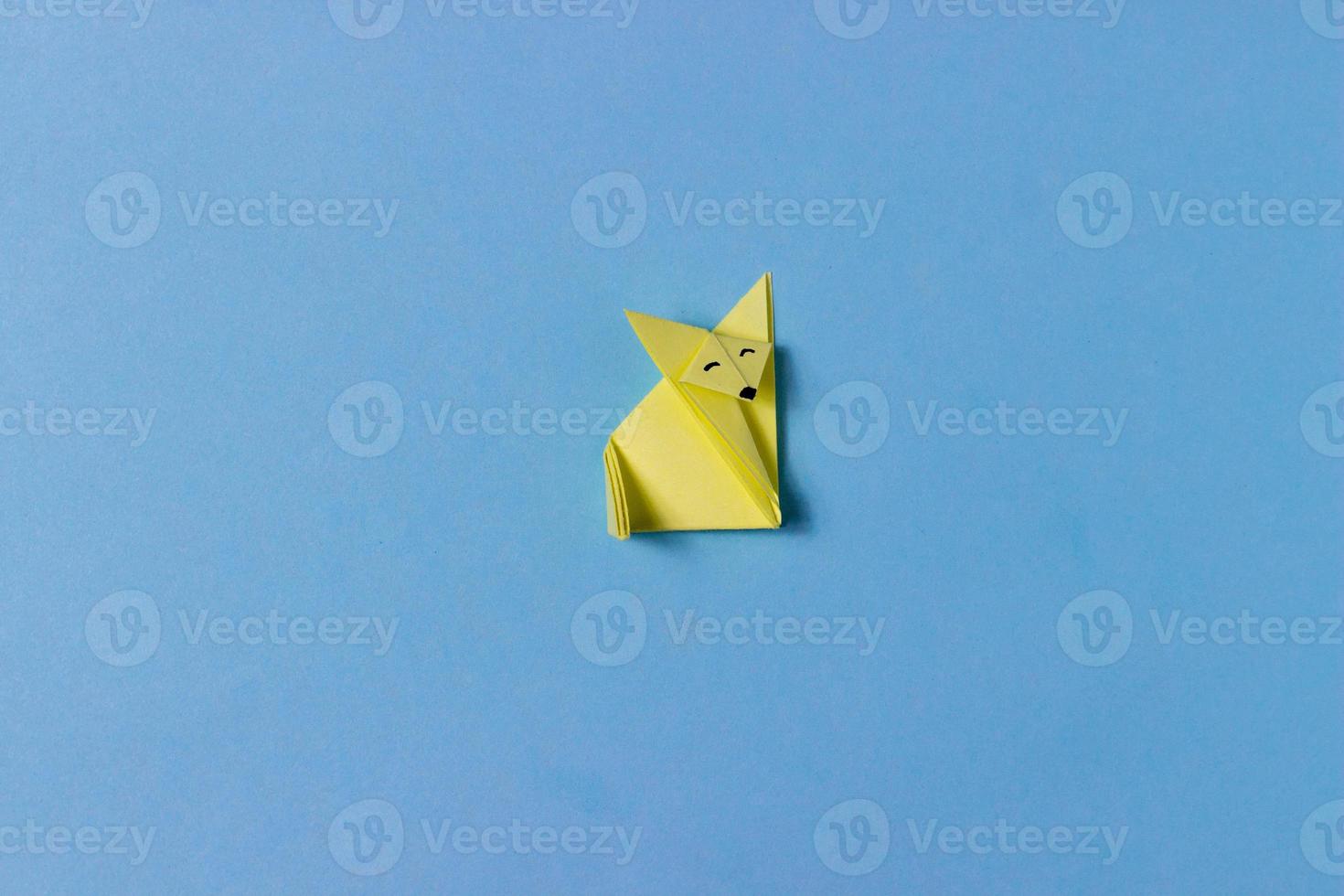 An origami fox is folded from yellow paper in the technique of origami with painted eyes, nose. In the middle of the blue background. Concept of love animals, hobbies, activities with children. photo