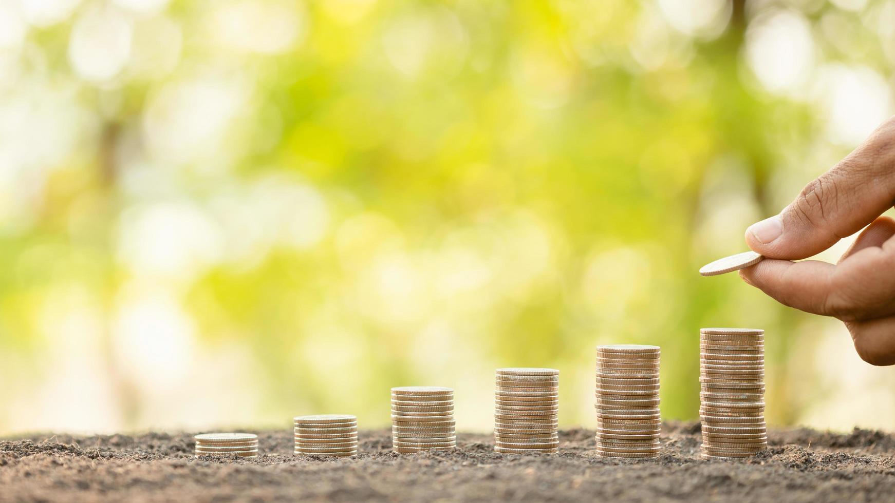 Hand putting coins in stack on wooden plank with green blur background. Money, Finance or Savings concept photo