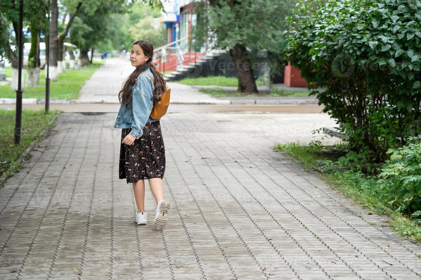 a girl in a dress and a denim jacket with an orange backpack on her shoulders goes to school and looks back. concept of back to school. Street photography. photo