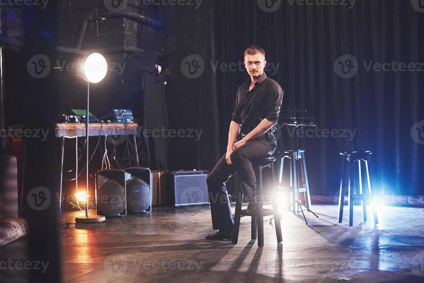 Magic look. Handsome young man in black clothes looking at camera while sitting on chair near the in dark room with light photo