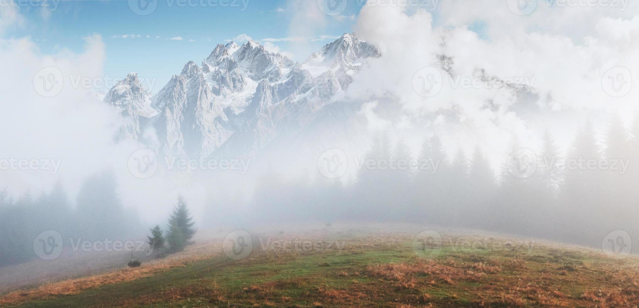 la niebla matutina se arrastra con restos sobre el bosque montañoso otoñal cubierto de hojas doradas. picos nevados de montañas majestuosas en el fondo foto