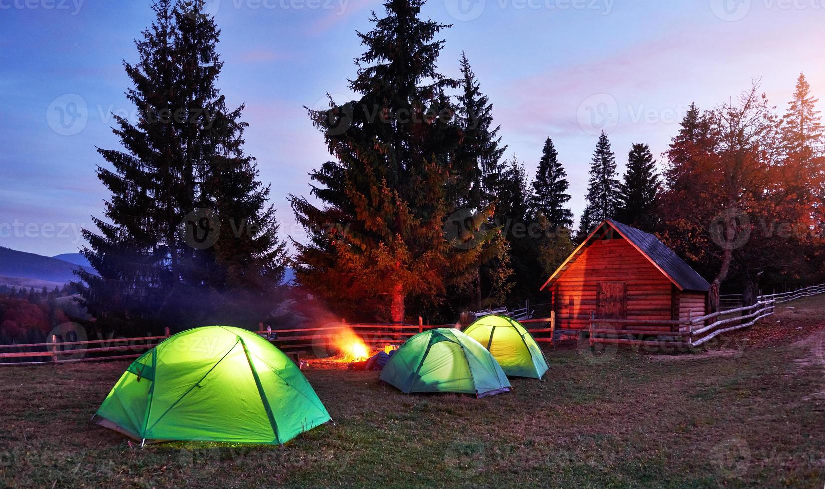 Night camping. Tourist have a rest at a campfire near illuminated tent and wooden house under amazing night sky full of stars and milky way. Astrophotography photo