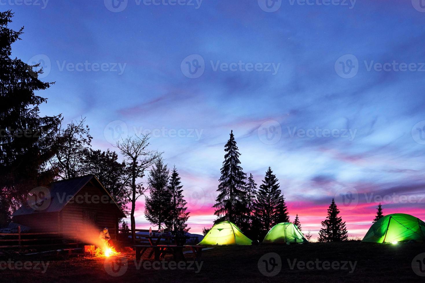 Night camping. Tourist have a rest at a campfire near illuminated tent and wooden house under amazing night sky full of stars and milky way photo