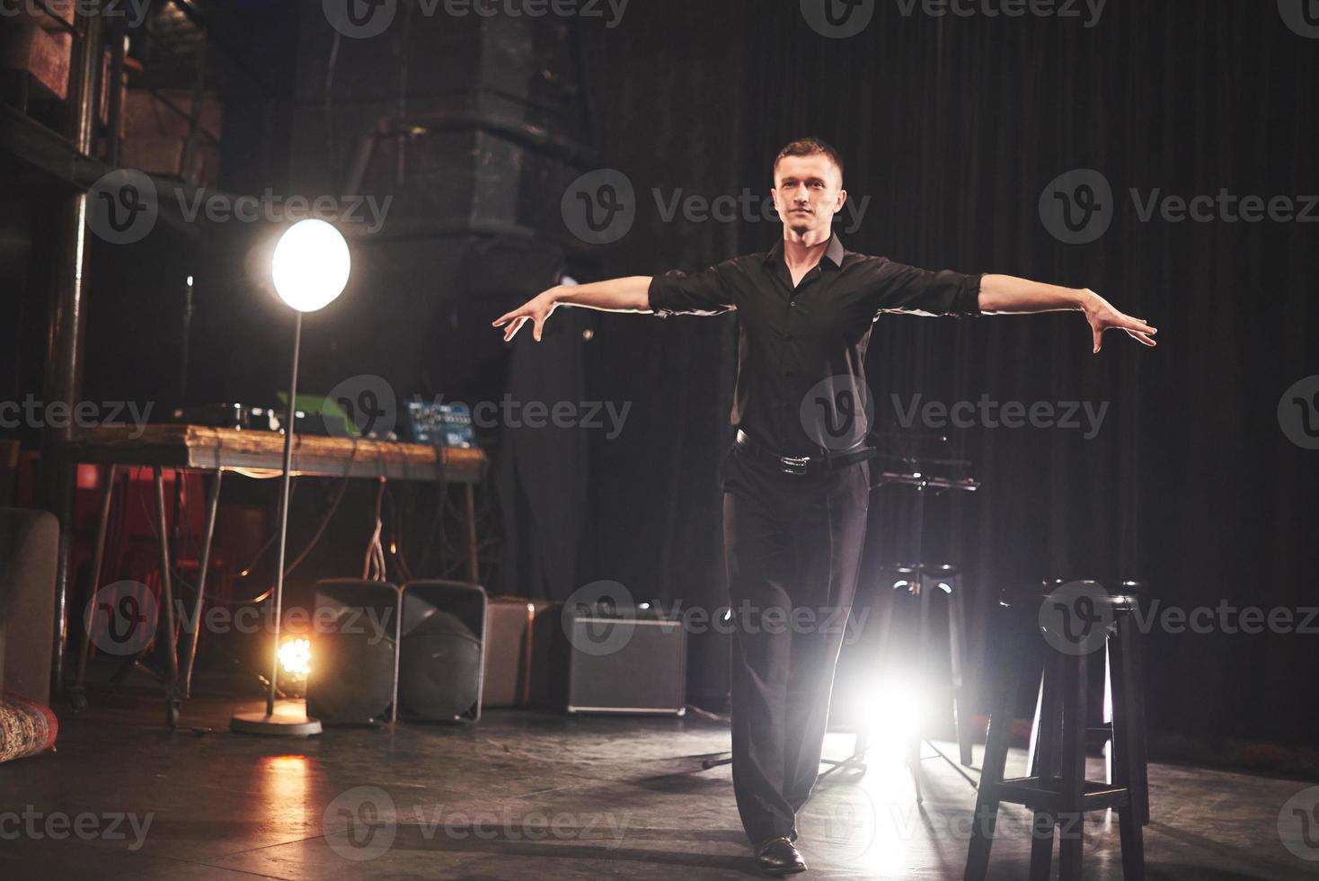 Magic look. Handsome young man in black clothes looking at camera while sitting on chair near the in dark room with light photo