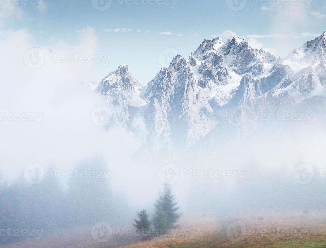 la niebla matutina se arrastra con restos sobre el bosque montañoso otoñal cubierto de hojas doradas. picos nevados de montañas majestuosas en el fondo foto