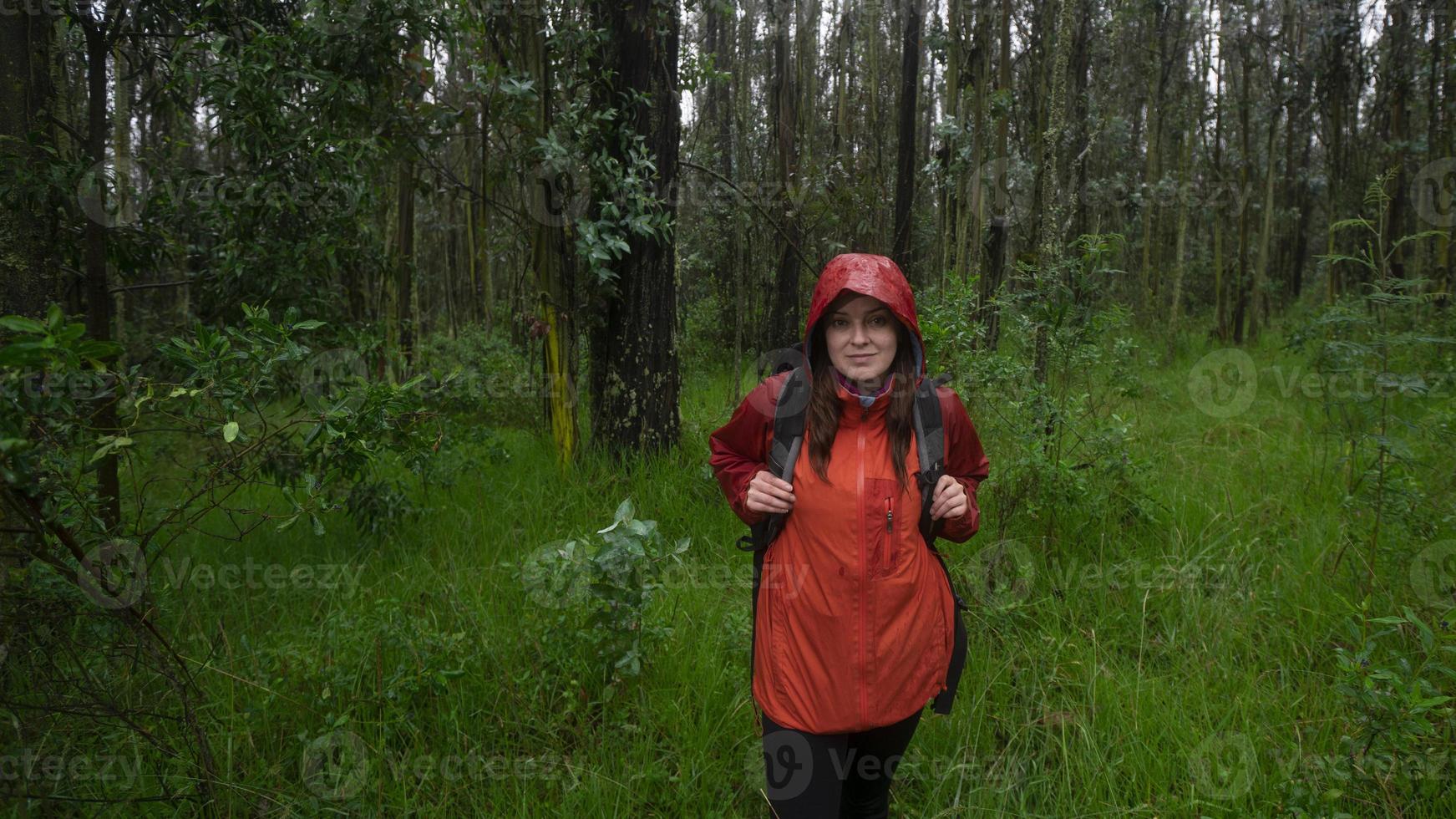 Hispanic woman with black backpack and red waterproof jacket walking on a muddy trail through a forest during the day photo