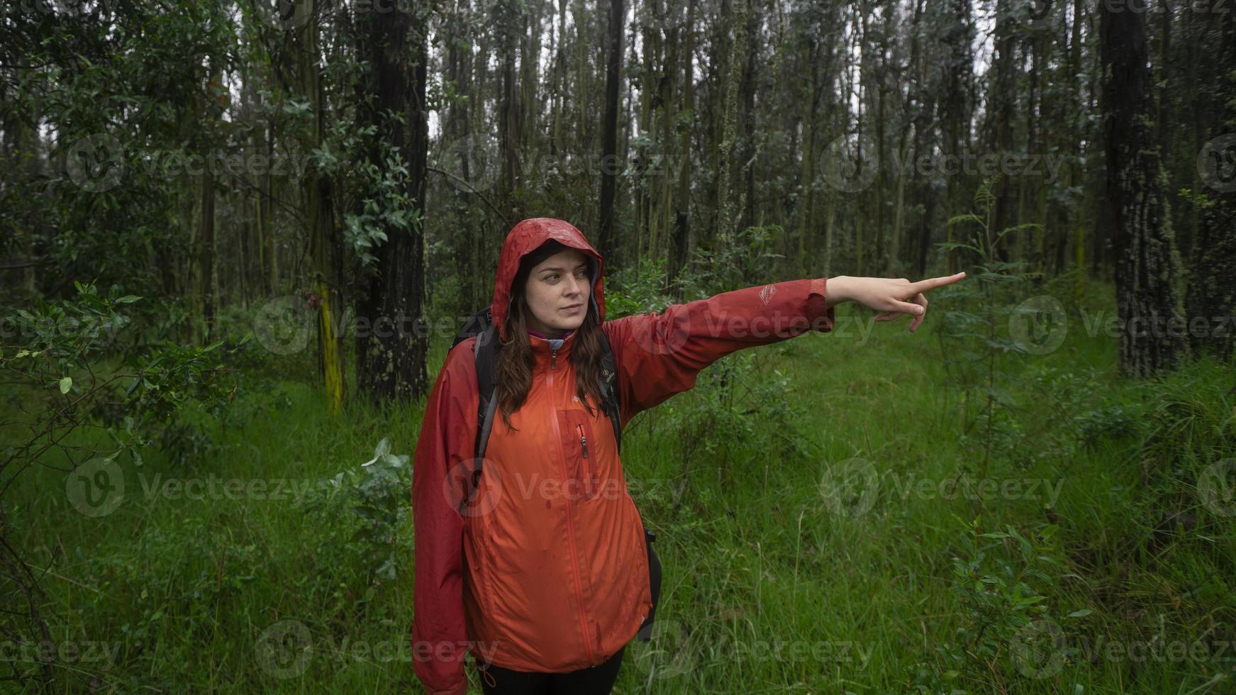 Front view of a Hispanic woman with a black backpack and a red waterproof jacket pointing the direction with her outstretched arm photo