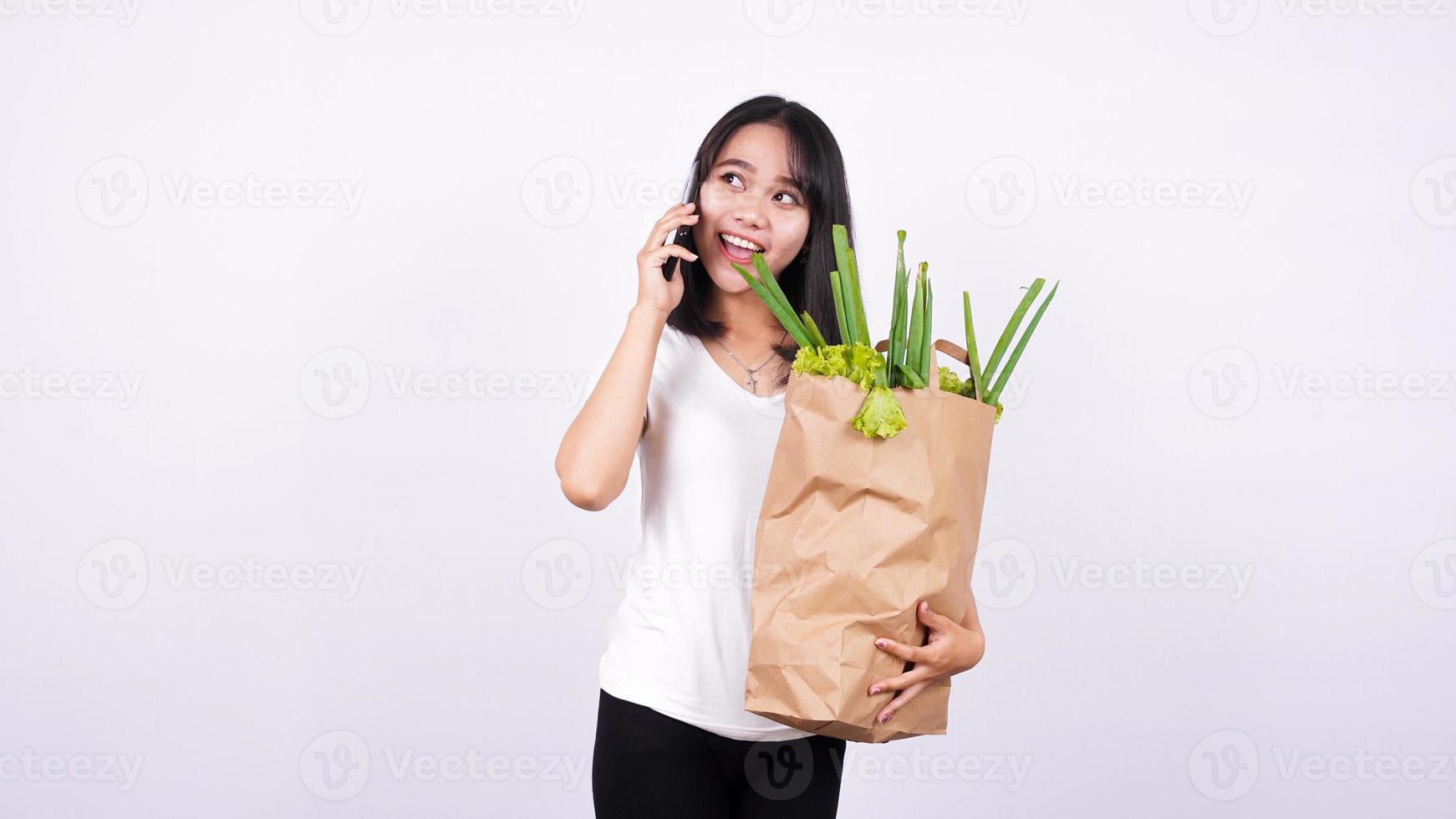 mujer asiática con una bolsa de papel de verduras frescas y hablando por teléfono con un fondo blanco aislado foto