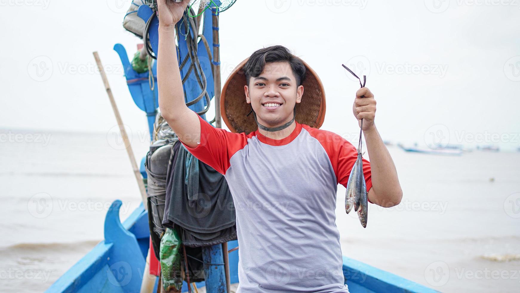 Happy young fisherman on the beach holding his catch fish and shows in front of his boat photo