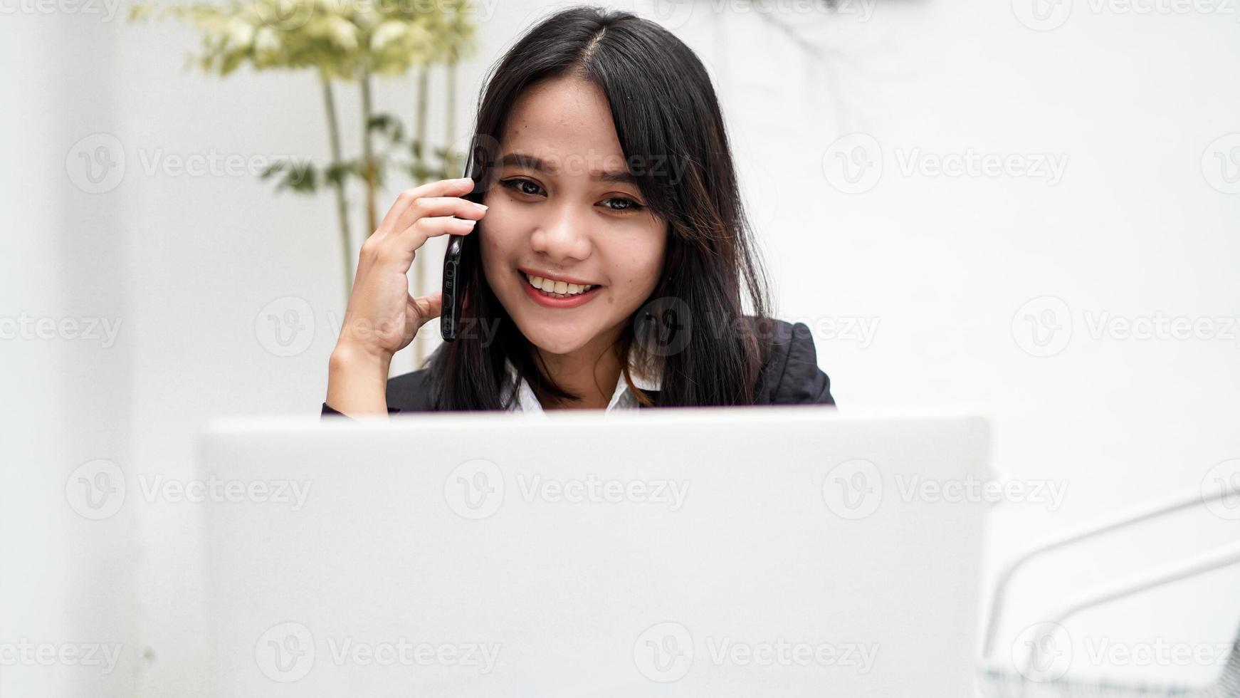 Asian young business woman working on computer and talking on smartphone in office photo