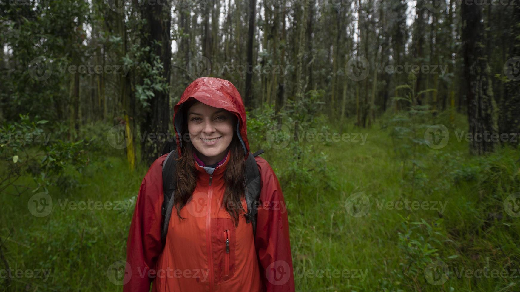 Hispanic woman with black backpack and red waterproof jacket walking on a muddy trail through a forest during the day photo