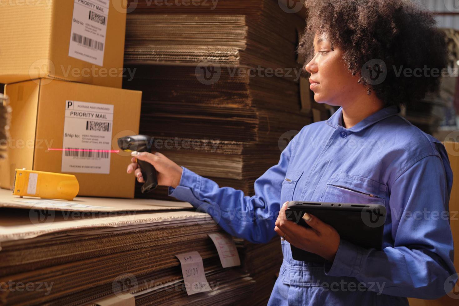 African American female worker in safety uniform using bar code scanner to check shipment orders at parcels warehouse, paper manufacture factory for the packing industry, logistic transport service. photo