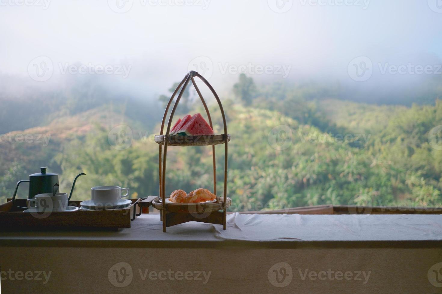 Pretty winter food set of natural resort breakfast meal, fresh watermelon, pie cup of coffee, and tea in kettle on wooden tray, eat in beautiful foggy mountain view, morning hill scenery landscape. photo