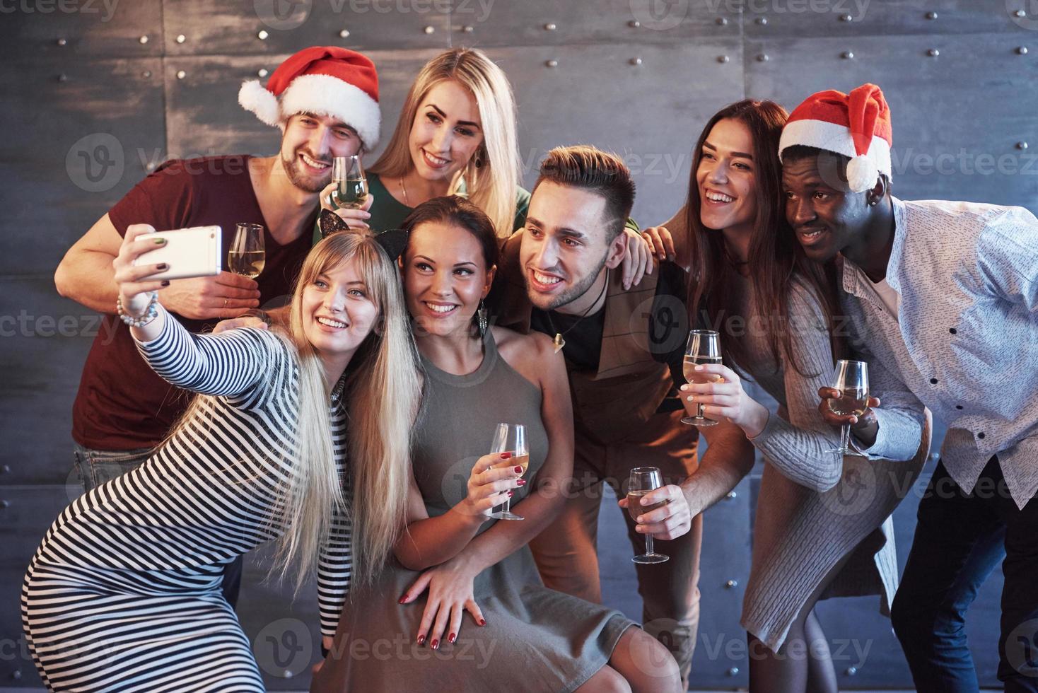 Agrupe a hermosos jóvenes haciendo selfie en la fiesta de año nuevo, mejores amigos niñas y niños divirtiéndose juntos, posando personas de estilo de vida emocional. sombreros de santa y copas de champán en sus manos foto