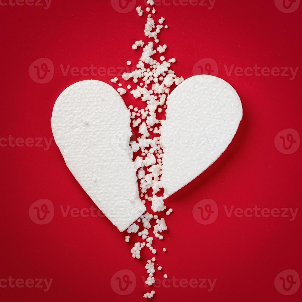 corazón de espuma de poliestireno roto blanco sobre un fondo de papel rojo. corazón desgarrado en dos partes con pequeñas partículas dispersas. concepto de amor infeliz foto