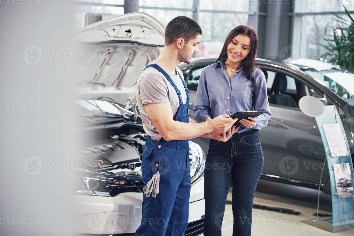 Beautiful young woman is talking to handsome car mechanic while repair a car in dealership photo