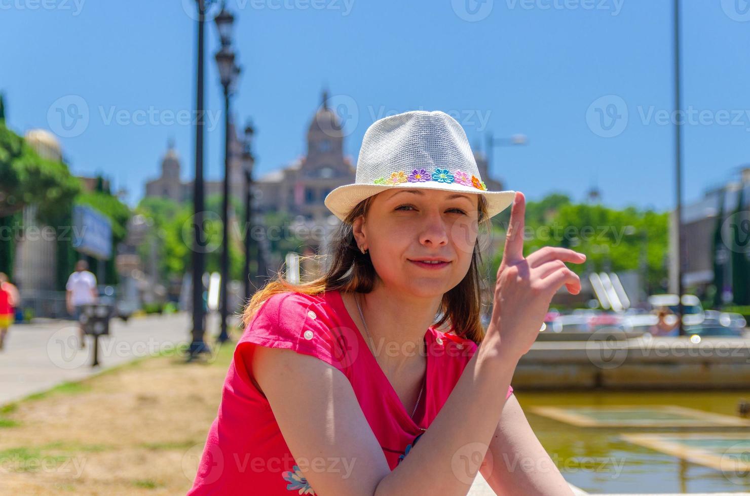 Young woman traveler with red dress and hat is sitting near fountain in Barcelona in sunny summer day photo