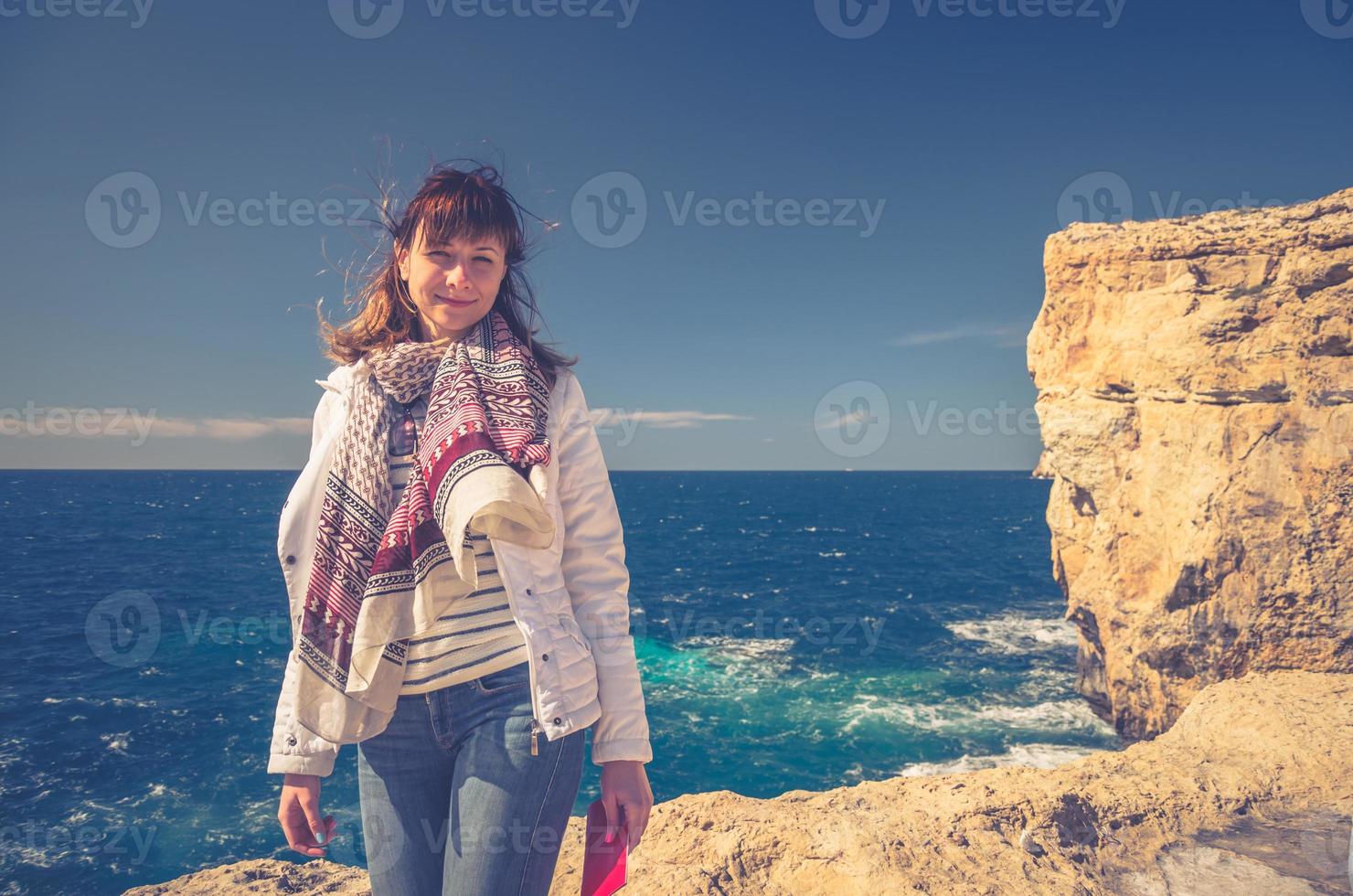 Young girl traveler with white jacket and headscarf looking at camera and smile on stone rocks of Dwejra Bay photo