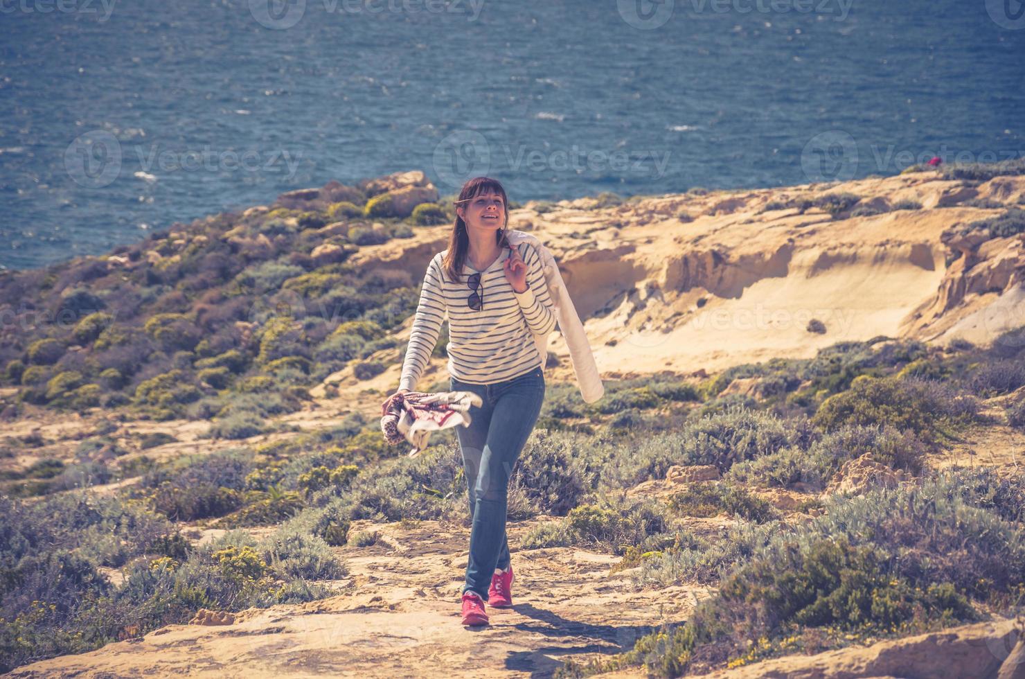 Young woman traveler with striped cardigan keep white jacket, smile and walking down coast cliffs of Dwejra Bay photo