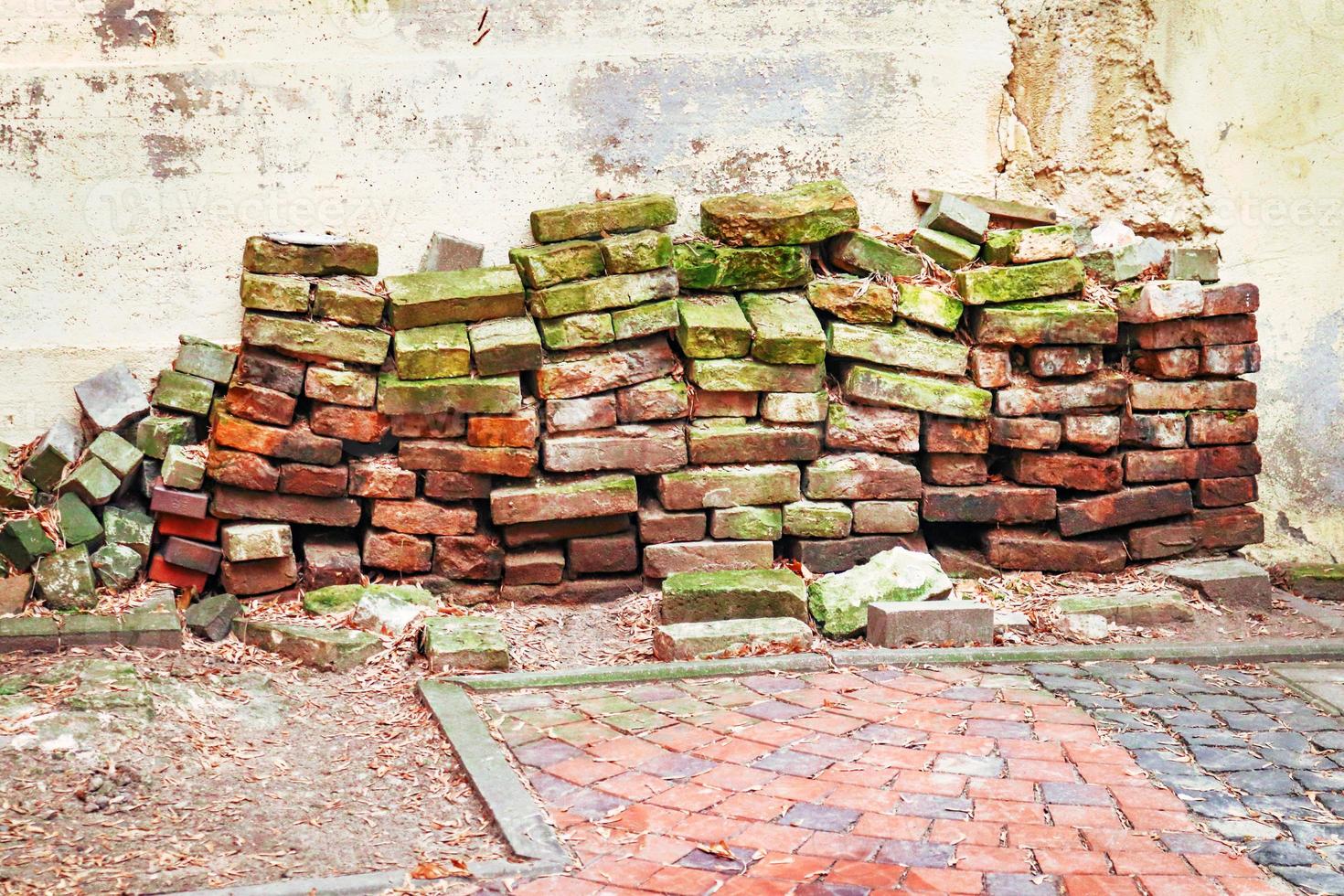 Aged red bricks stacked in yard on red pavement along white wall photo
