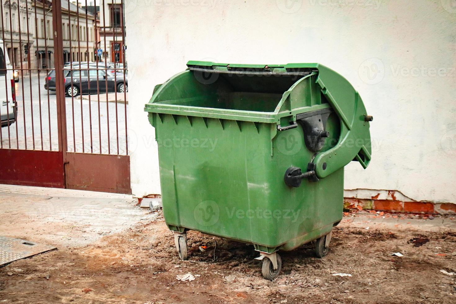 Green garbage container with open cover standing outdoors on dry sandy ground near light wall with red metal gates photo