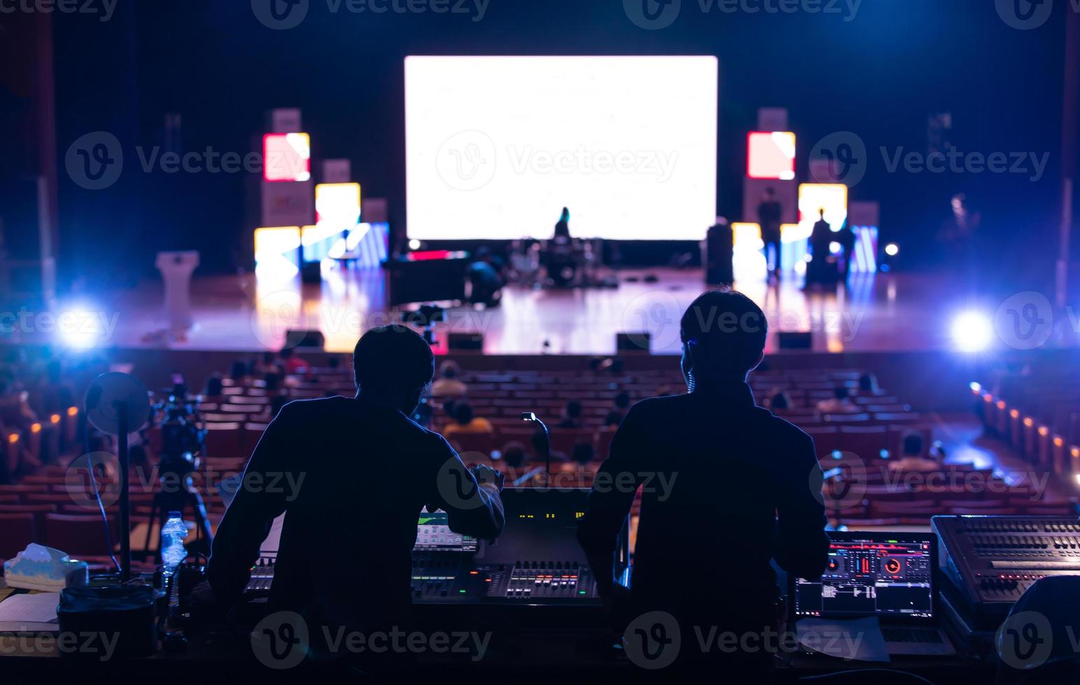 equipo de ingenieros de sonido trabajando en la preparación de mezcladores y equipos frente al escenario del concierto con pantalla grande borrosa. foto