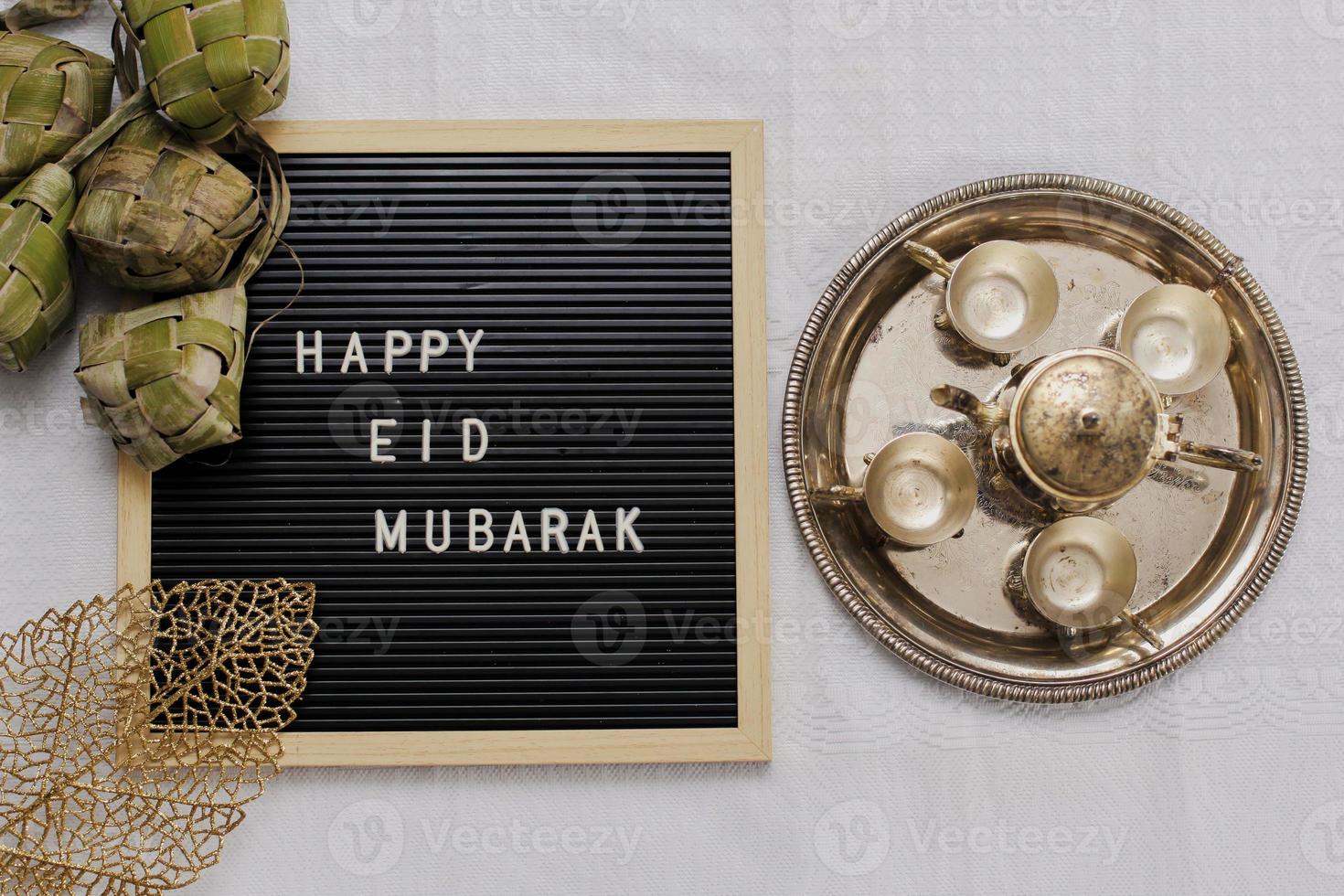 Top view of letter board says Happy Eid Mubarak and traditional food during lebaran celebration called ketupat with set golden teapot and cups photo