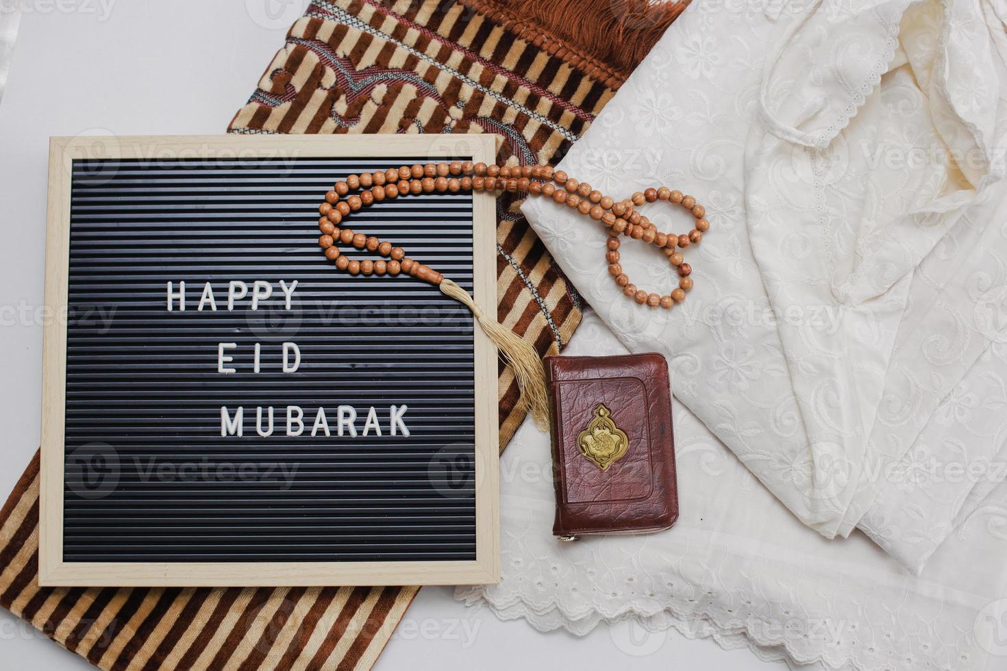 falt lay de vestido musulmán llamado mukena y cuentas de oración con el libro sagrado de al quran y tablero de letras dice feliz eid mubarak en la alfombra de oración. hay una letra árabe que significa el libro sagrado foto