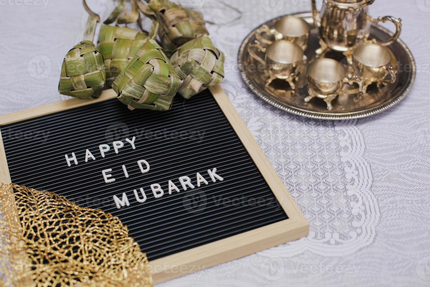 Close up of letter board says Happy Eid Mubarak and traditional food during lebaran celebration called ketupat with set golden teapot and cups photo