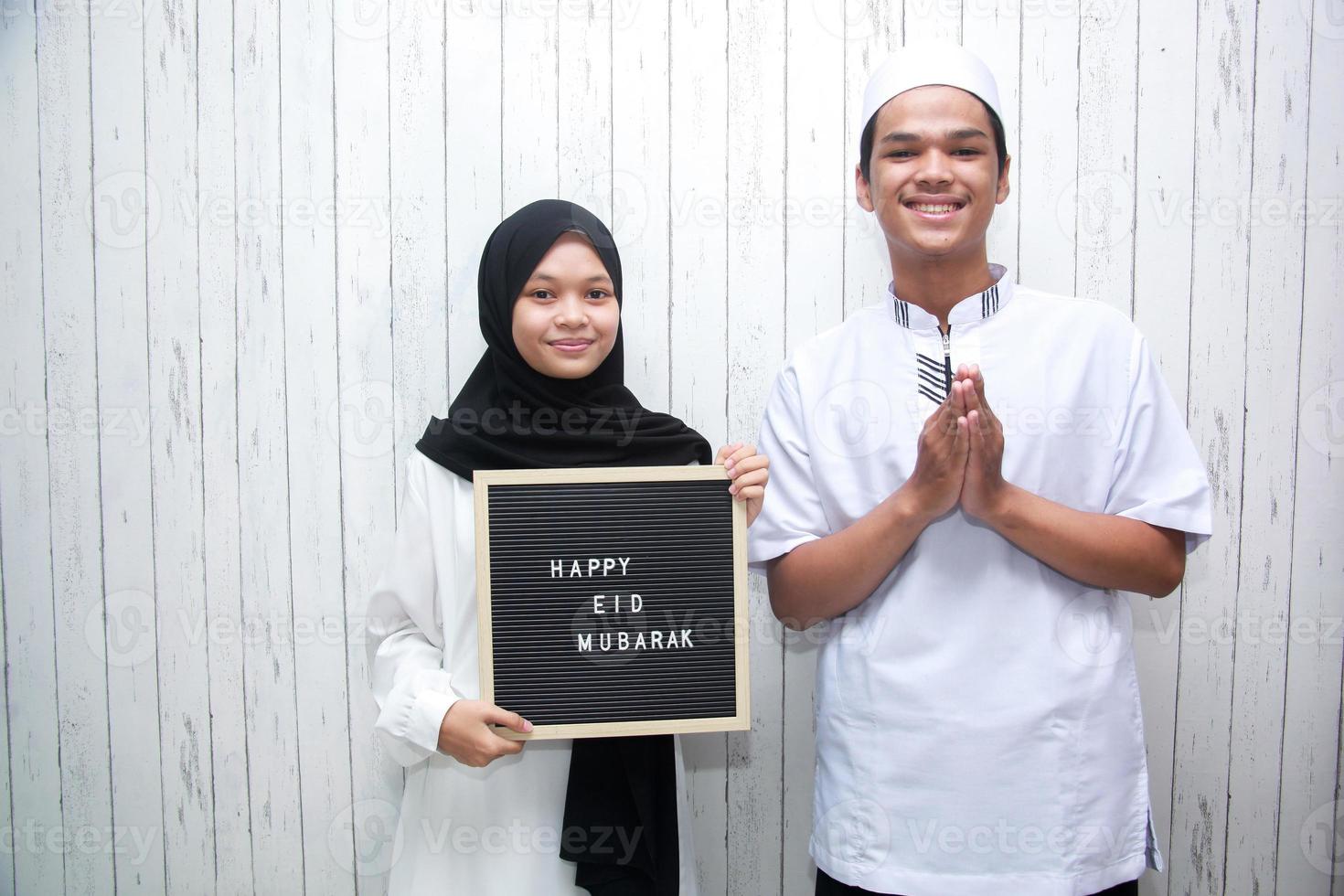 joven pareja musulmana haciendo una pose de saludo y sosteniendo un tablero de cartas dice feliz eid mubarak foto