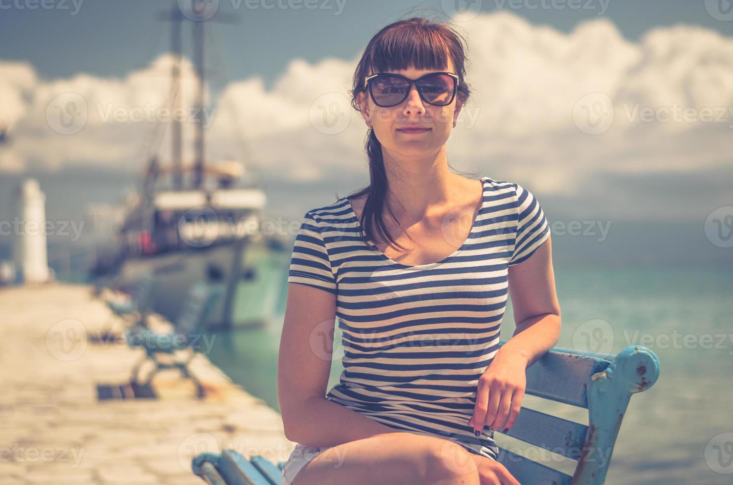 hermosa joven con camiseta a rayas y gafas de sol sonríe y se sienta en un banco en el muelle foto