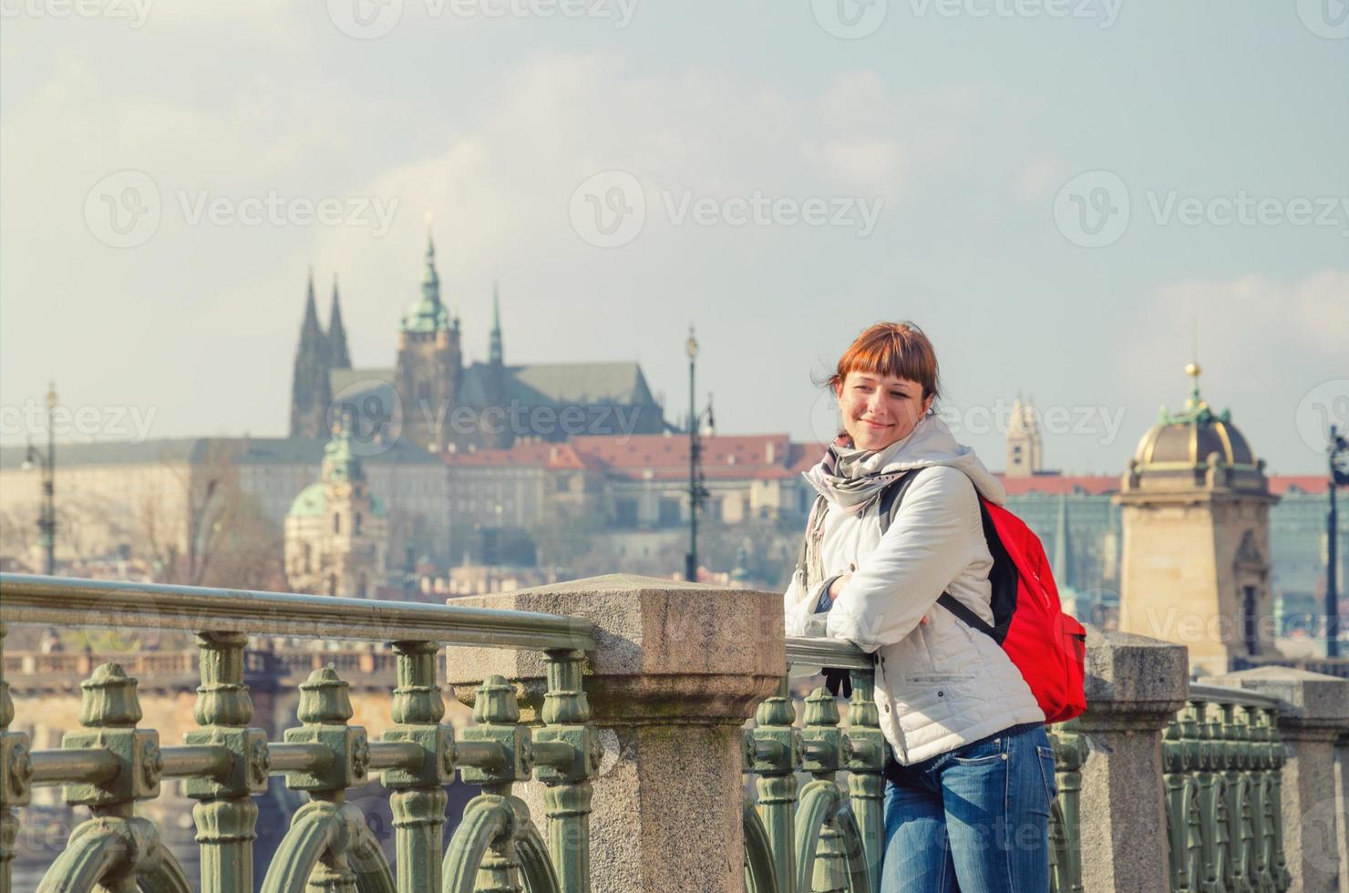 Young girl tourist with white jacket and red backpack looking away and posing near fence of Vltava river photo