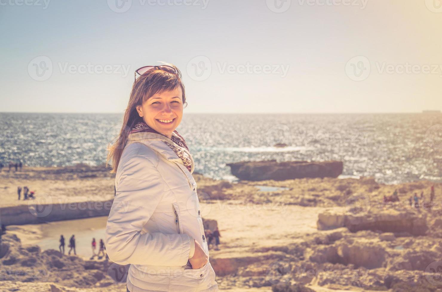 joven viajera con chaqueta blanca y gafas de sol mirando a la cámara y sonriendo en la costa de la bahía de dwejra foto