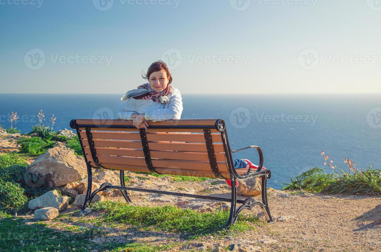 Young beautiful girl traveler with white jacket looking at camera, sit and smile on bench at Dingli Cliffs photo