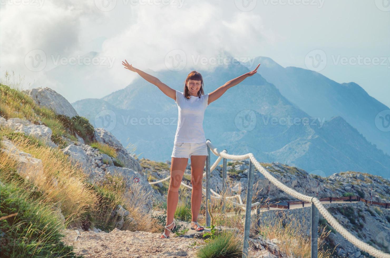 joven viajera caucásica con camiseta blanca y pantalones cortos posando, levanta la mano y sonríe en el camino foto