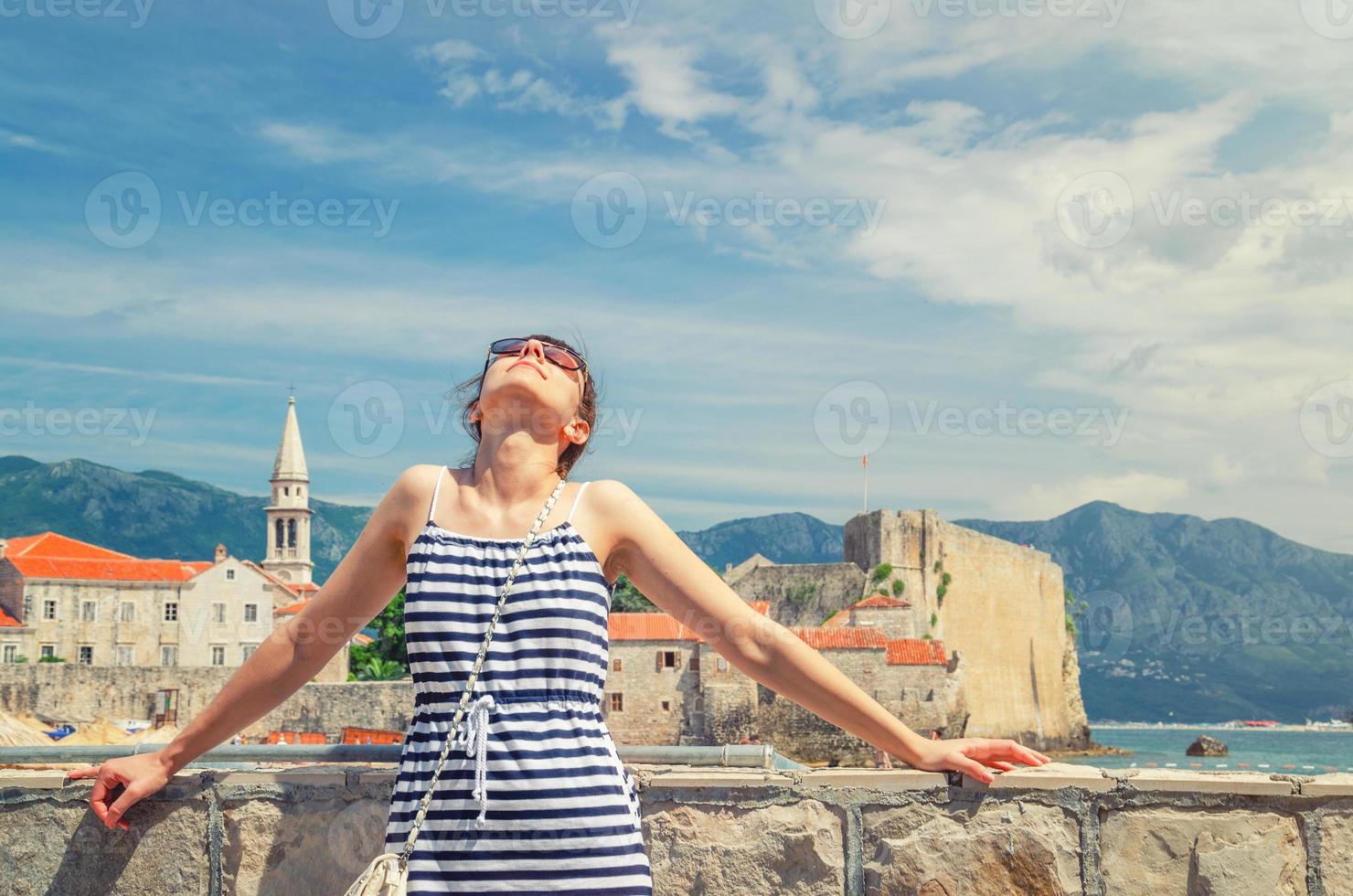 joven hermosa chica viajera con vestido a rayas y gafas de sol mirando hacia arriba y posando cerca del mar adriático foto