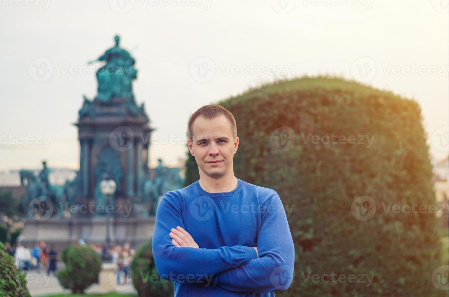 Young man traveler looking at camera, smile and posing, green bush and Empress Maria Theresia monument photo