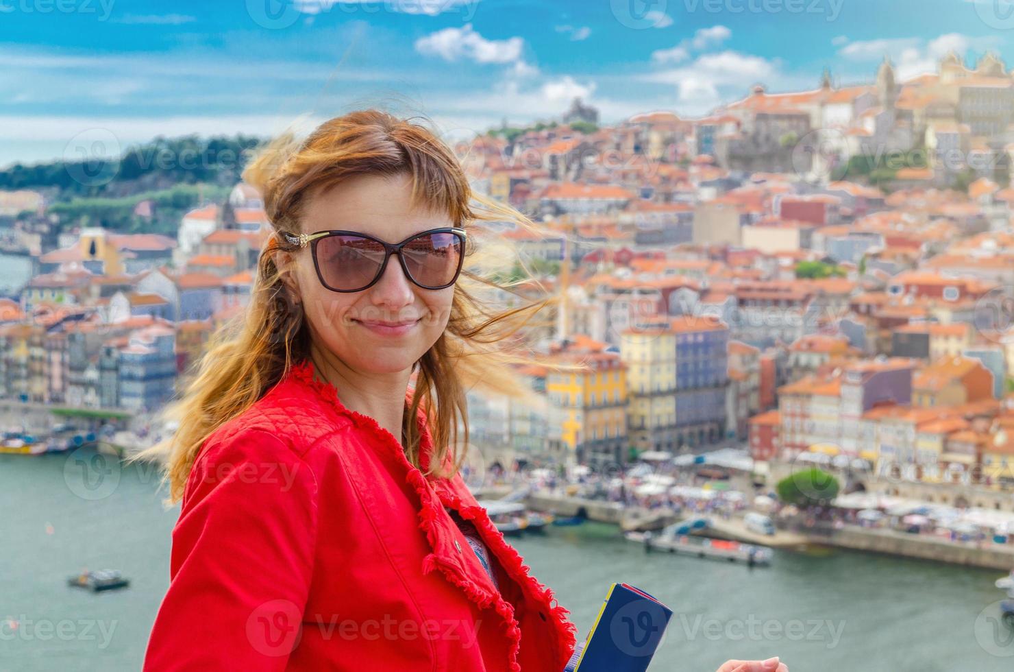 Young woman traveler with sunglasses and red jacket looking at camera, smiling and posing in front of Porto Oporto city photo