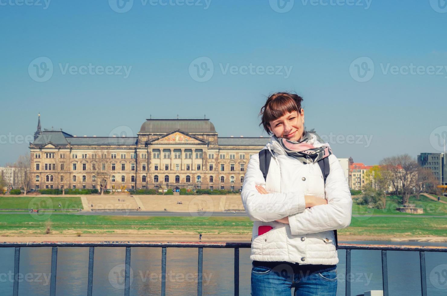 joven viajera con chaqueta blanca mirando a la cámara y sonriendo, río elba con edificios antiguos casas en la ciudad de dresden foto