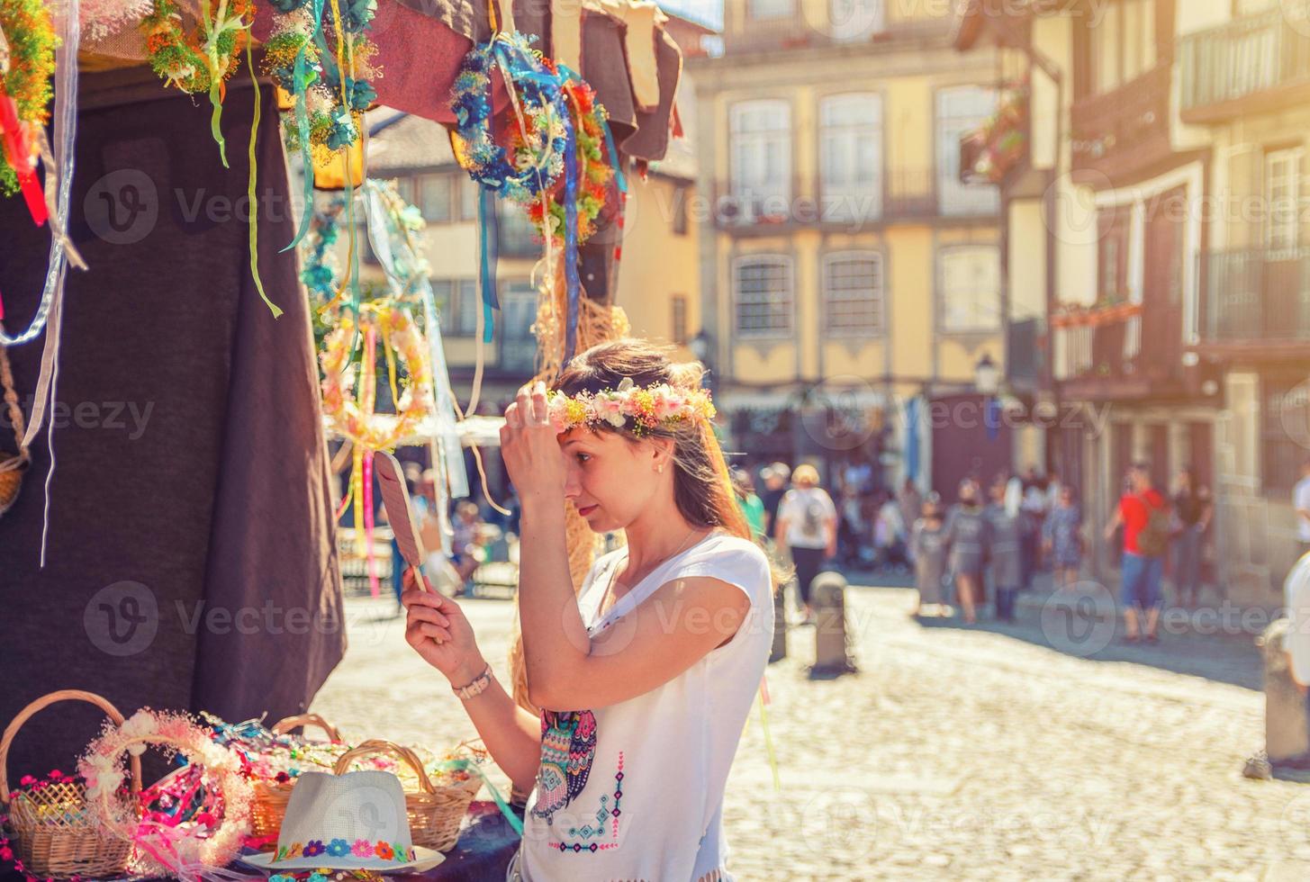 Young woman traveler trying on flower crown wreath and looking at hand mirror near souvenir booth stand photo