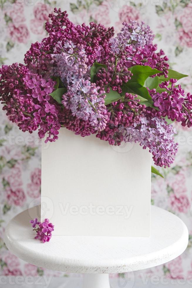 White sheet in beautiful blooming lilac on a white stand on a floral background. Greeting card, place for text, mock up photo