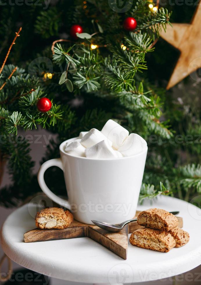 café en una taza blanca con malvaviscos. café festivo matutino con galletas de almendra cantuccini italianas tradicionales. una taza de café sobre un fondo de ramas de abeto verde sobre un soporte blanco. foto