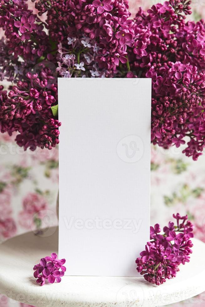 White sheet in beautiful blooming lilac on a white stand on a floral background. Greeting card, place for text, mock up photo