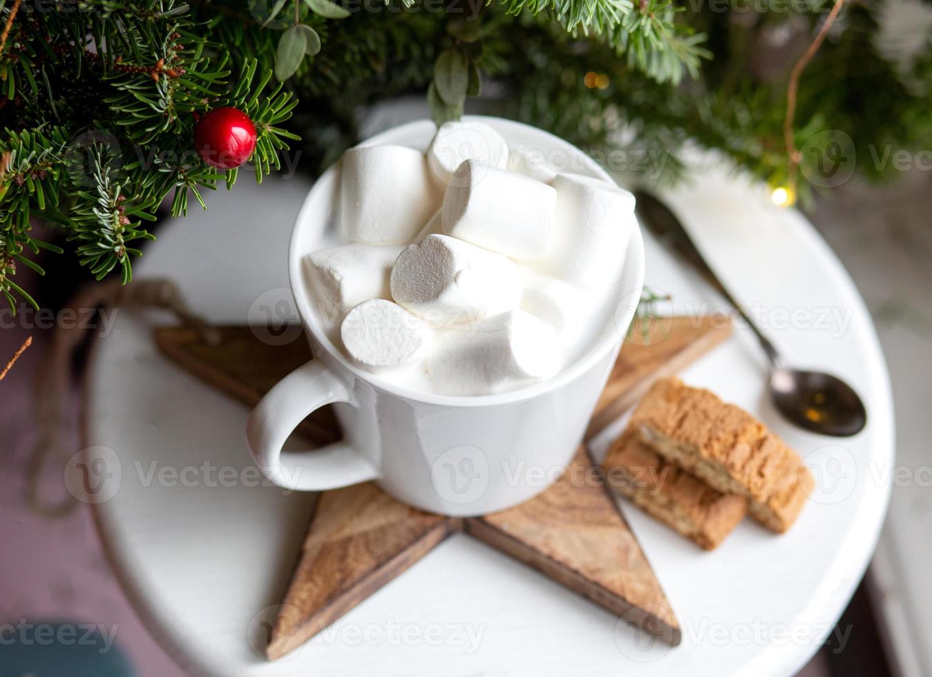 café en una taza blanca con malvaviscos. café festivo matutino con galletas de almendra cantuccini italianas tradicionales. una taza de café sobre un fondo de ramas de abeto verde sobre un soporte blanco. foto