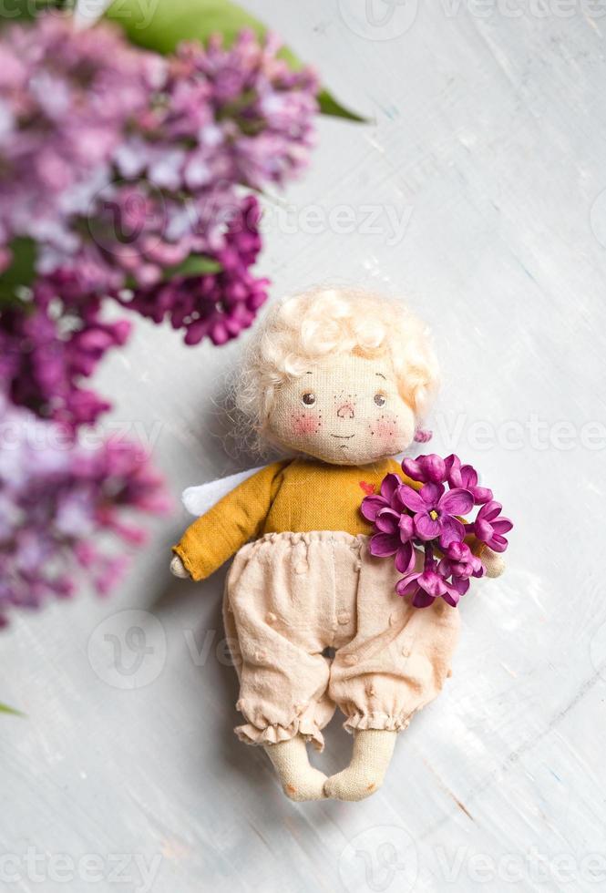 angelito de pelo dorado en las flores lilas azules, rosas, violetas y violetas. juguete hecho a mano en colores violeta lila. tarjeta de felicitación. foto