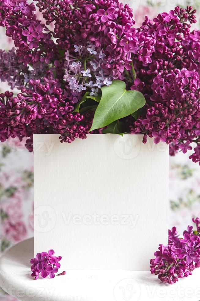 White sheet in beautiful blooming lilac on a white stand on a floral background. Greeting card, place for text, mock up photo