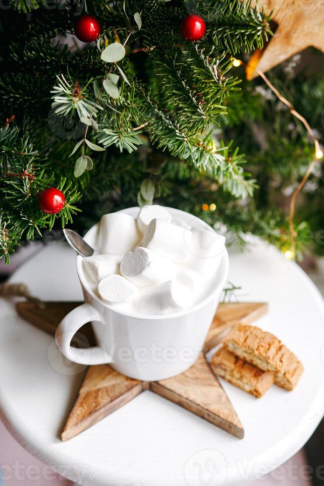 Coffee in a white cup with marshmallows. Morning festive coffee with traditional Italian cantuccini almond cookies. A cup of coffee on a background of green fir branches on a white stand. photo