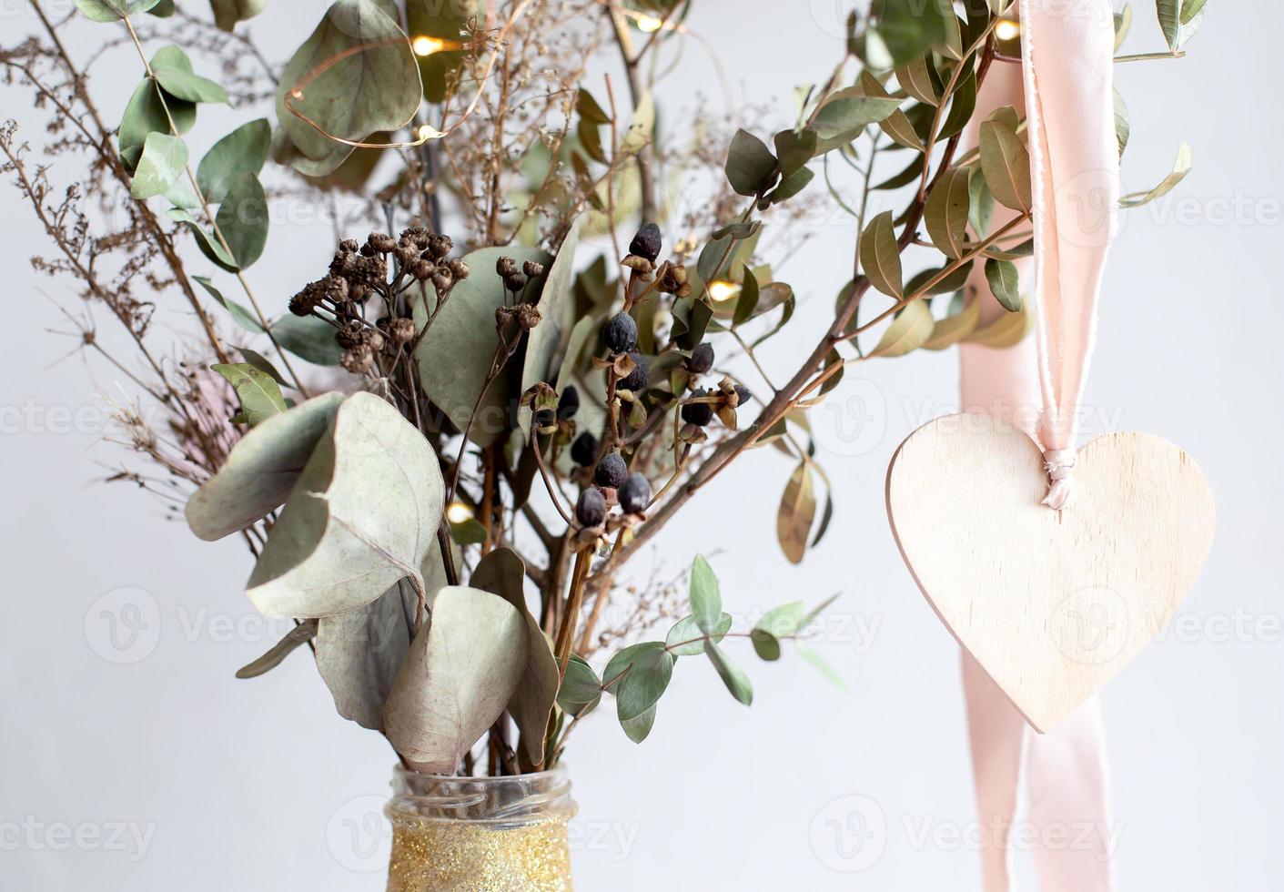 Light wooden heart pendant on a pink silk ribbon on a background of a golden shiny glittered vase with dried eucalyptus branches. photo