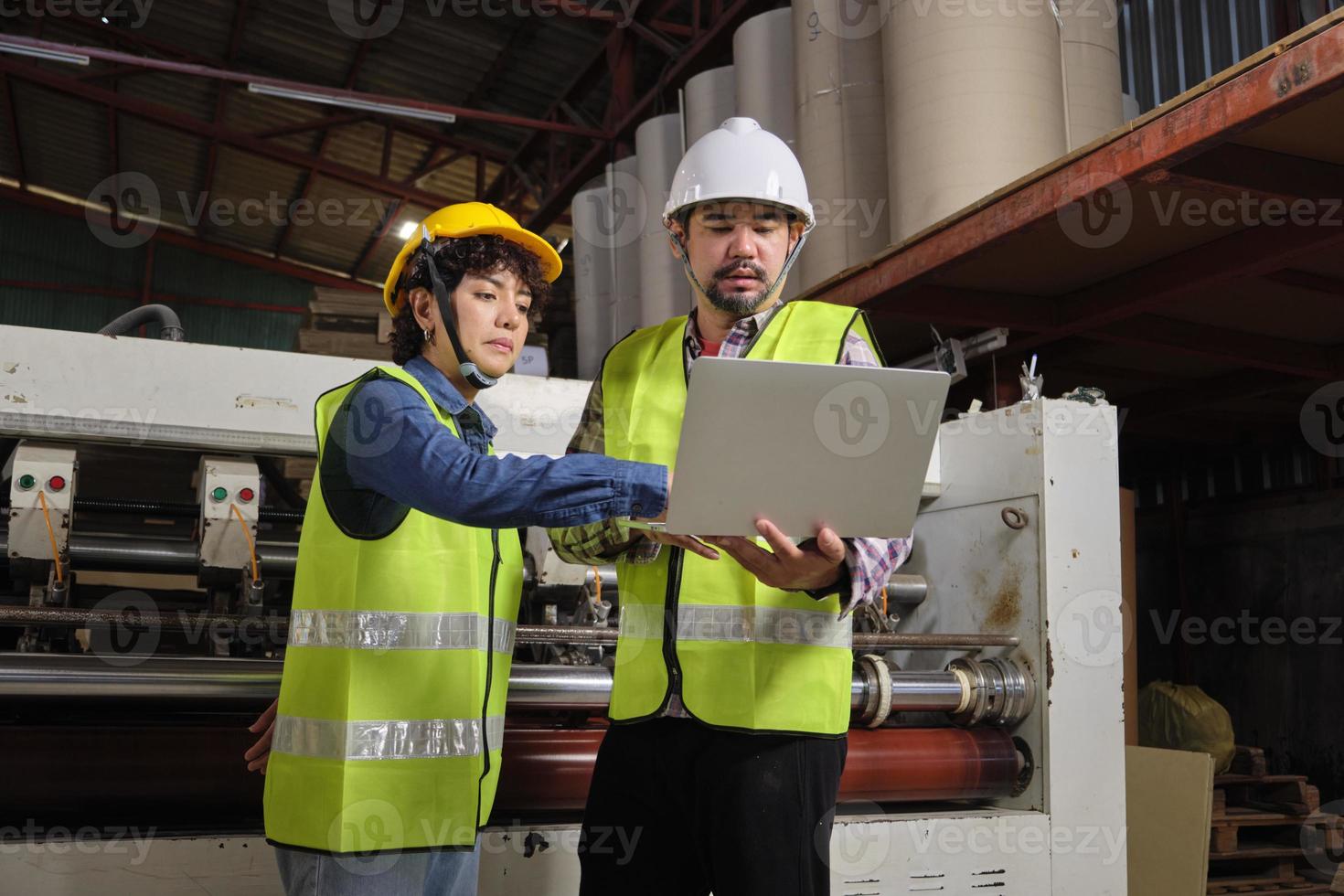 Safety uniform workers and industrial engineers with hardhat use laptop computer to check and control machines. Two professionals work in paper manufacturing factory, maintain production equipment. photo