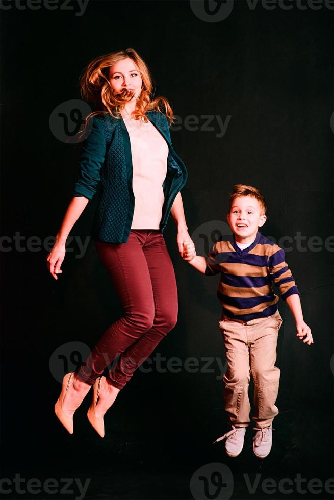 portrait of stylish cute little boy with beautiful mom jumping together on dark background in photo studio